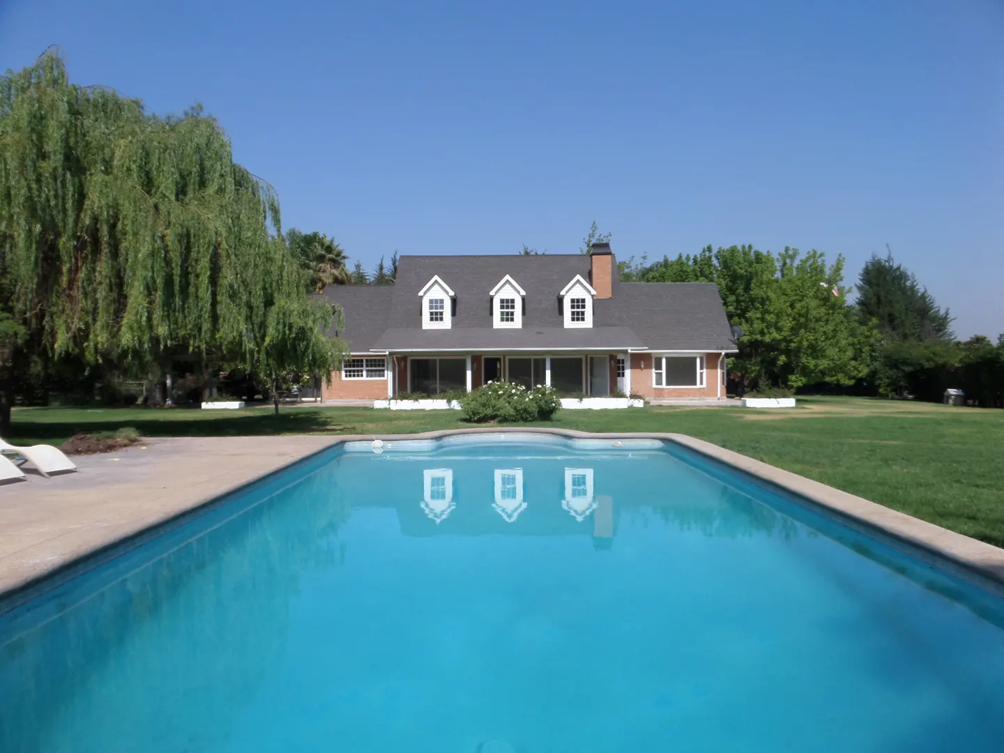 A bright blue swimming pool reflects a brick house with a gray roof and white dormers on a sunny day.