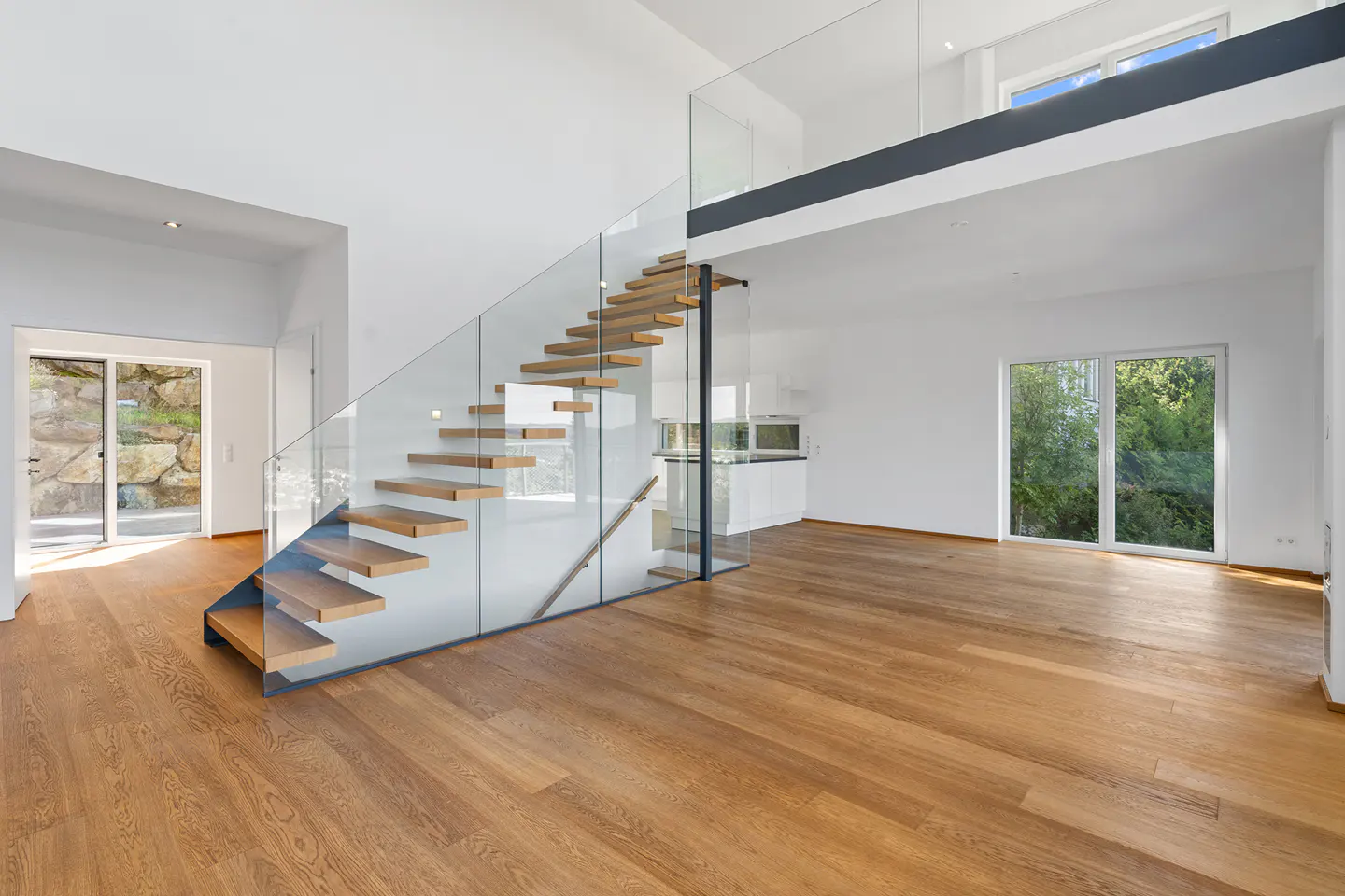 Bright, modern interior with wood floors, white walls, and floating wood stairs with glass railing. A loft overlooks the open space.