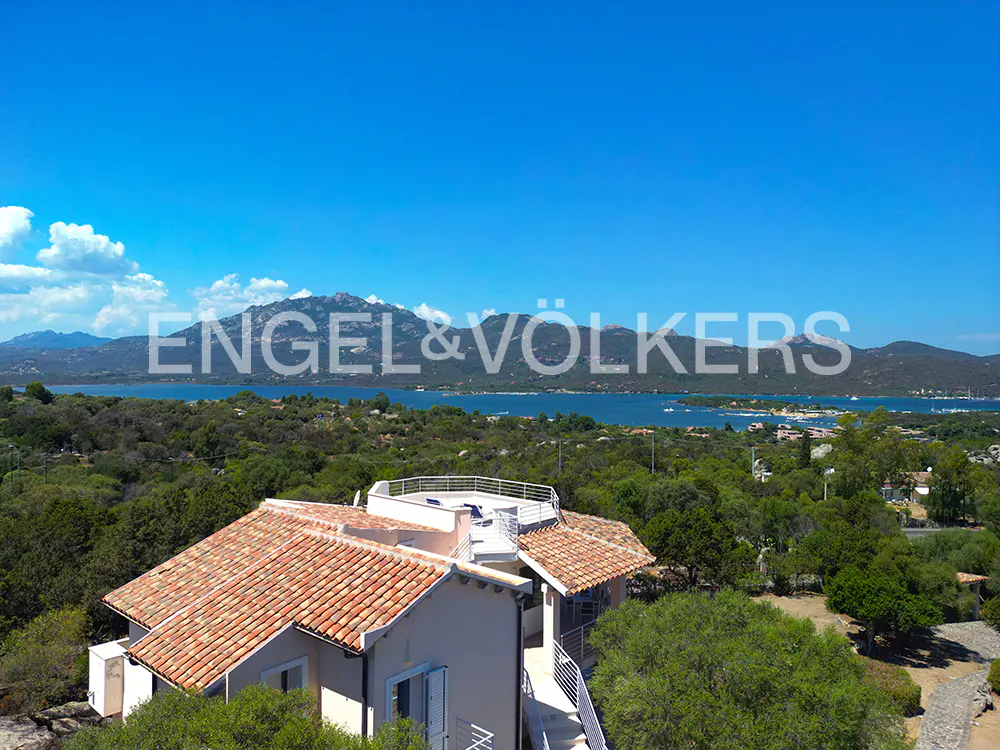 Aerial view of a villa with a terracotta roof, surrounded by lush greenery, overlooking a bay and distant mountains.