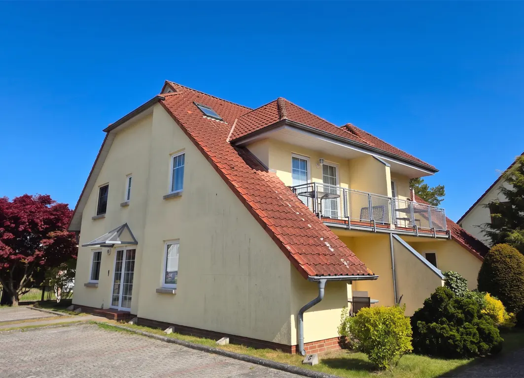 Exterior view of a two-story yellow house with a red tile roof and a balcony, set against a clear blue sky.