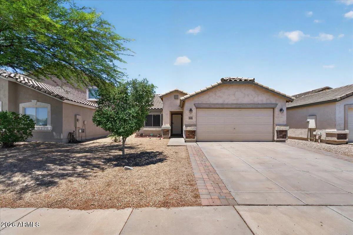 Single-story beige stucco house with a two-car garage, concrete driveway, and desert landscaping under a blue sky.