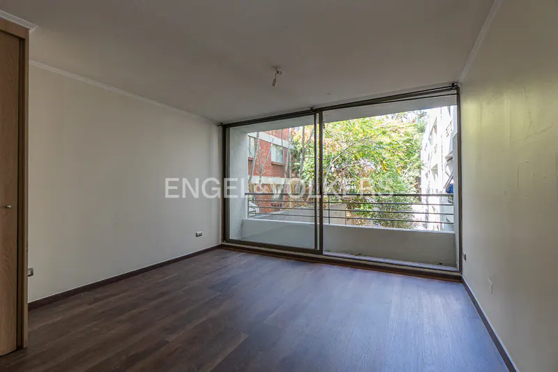 Empty room with dark wood floors and white walls. Large sliding glass doors lead to a balcony with trees and buildings visible.