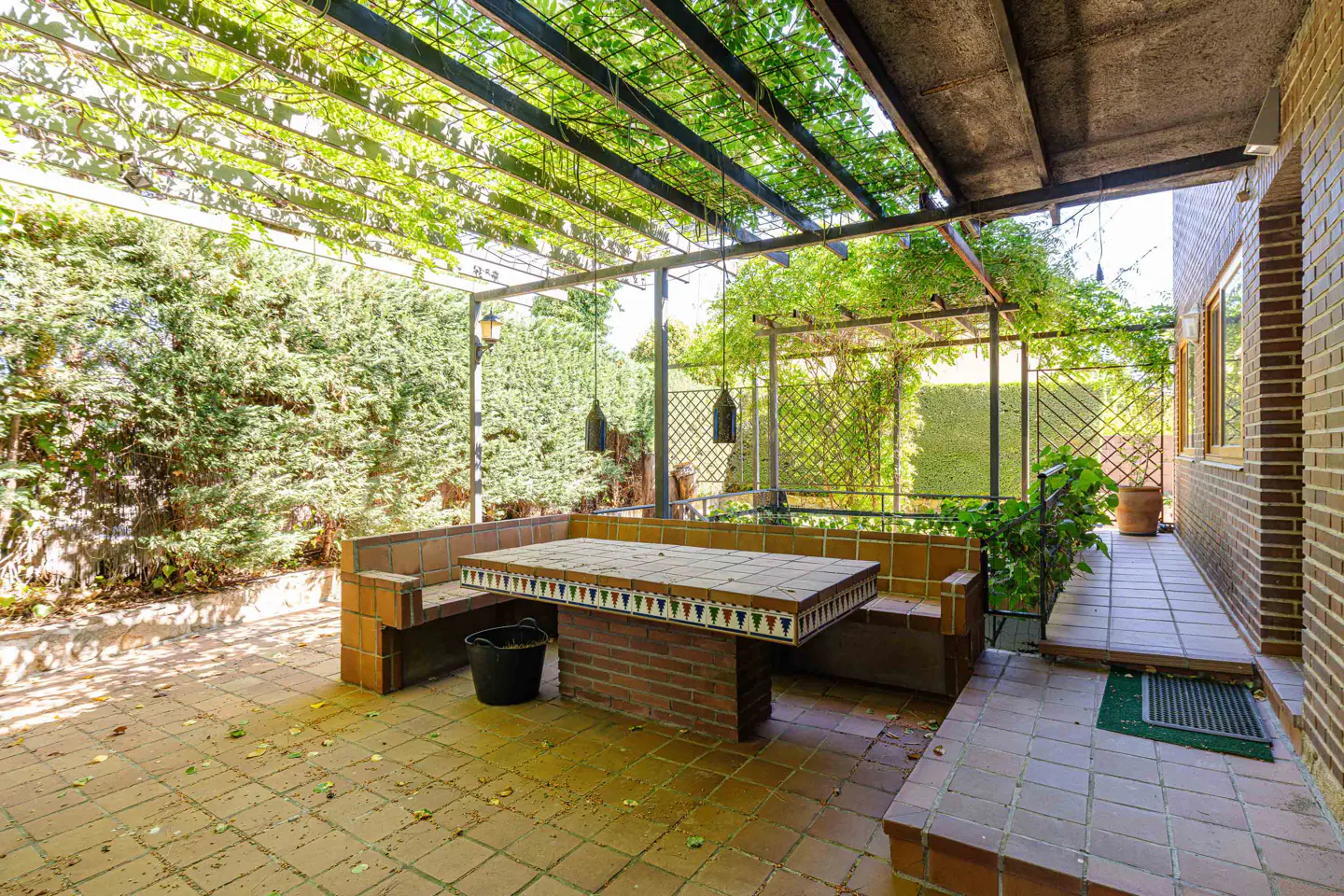 Outdoor patio with a tiled table and built-in bench under a vine-covered pergola, surrounded by lush greenery and brick.