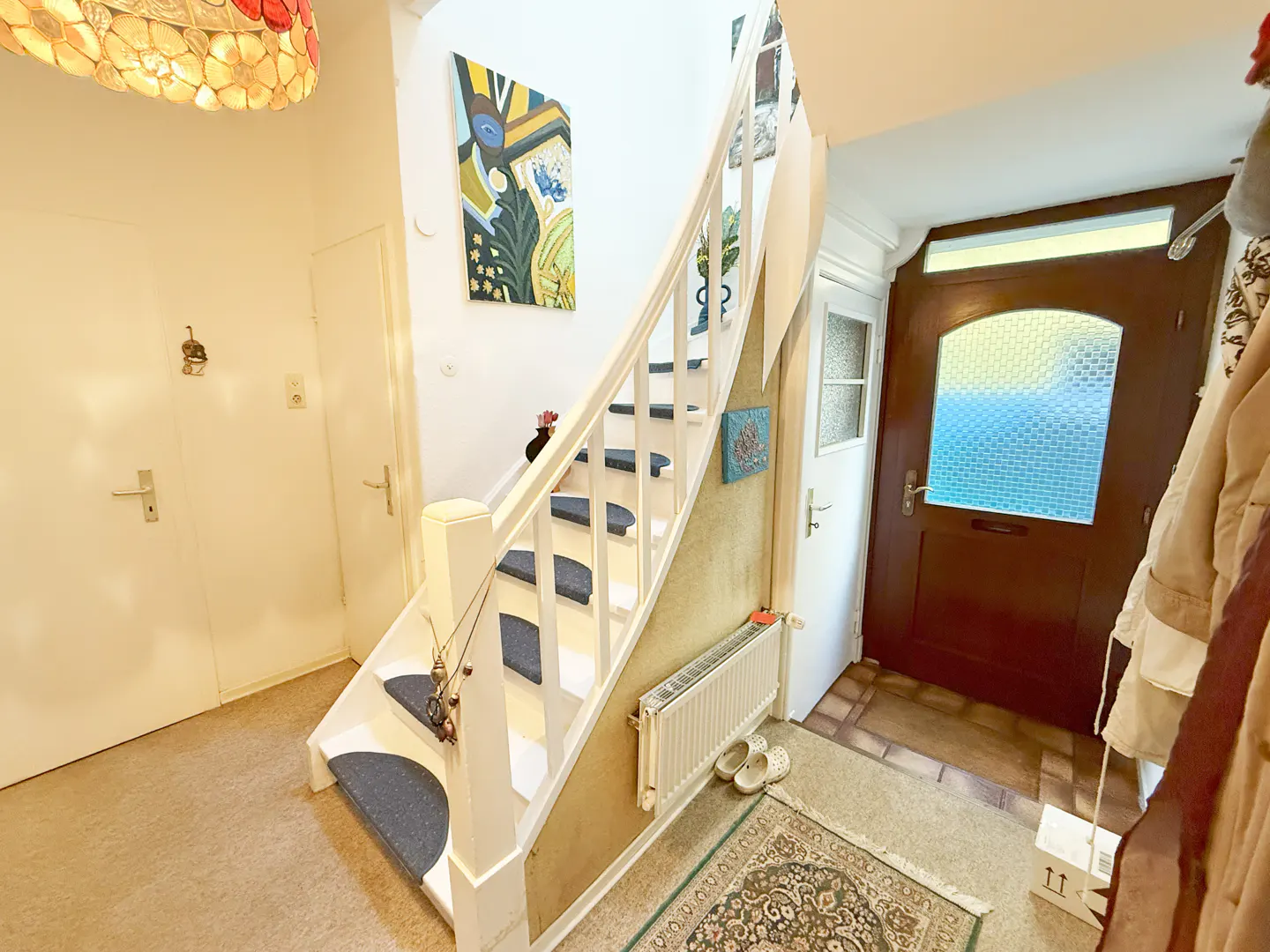 A bright foyer with white walls, a curved staircase with blue carpet, and a dark wood front door with a textured glass window.
