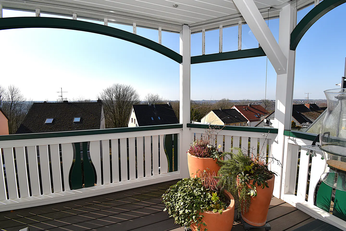 Balcony view with potted plants. White railing with green trim overlooks houses under a blue sky.