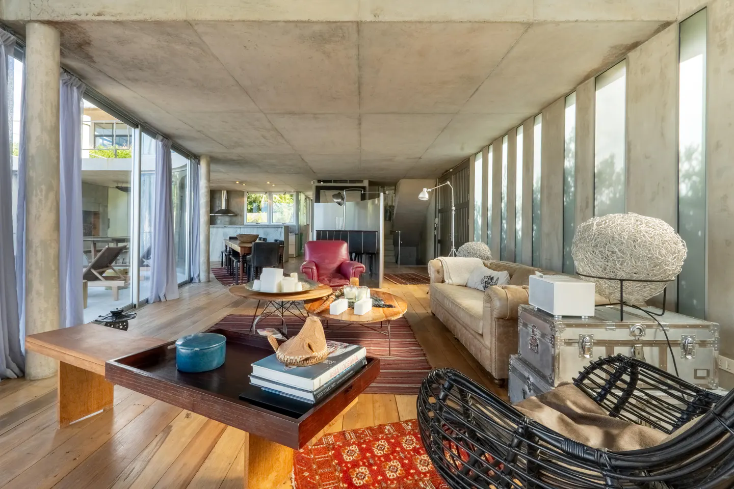 Open-concept living room with concrete walls, wood floors, and large windows. Furnished with a beige sofa, red chair, and wicker furniture.