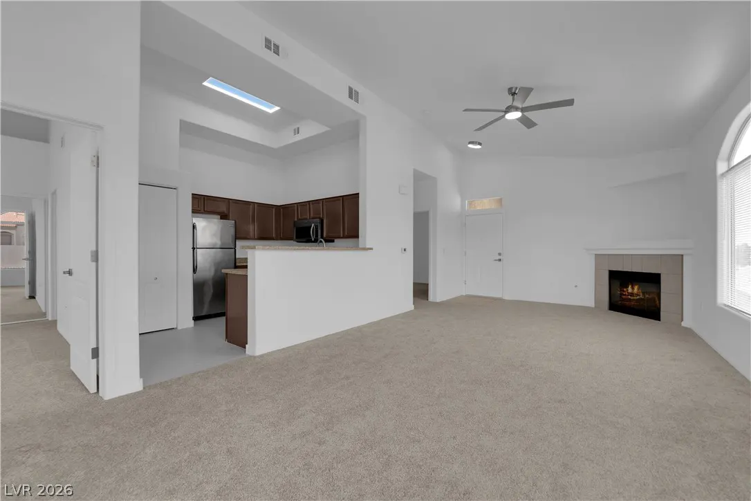 Open-concept living space with white walls, beige carpet, and a fireplace. Kitchen with brown cabinets and stainless steel appliances.