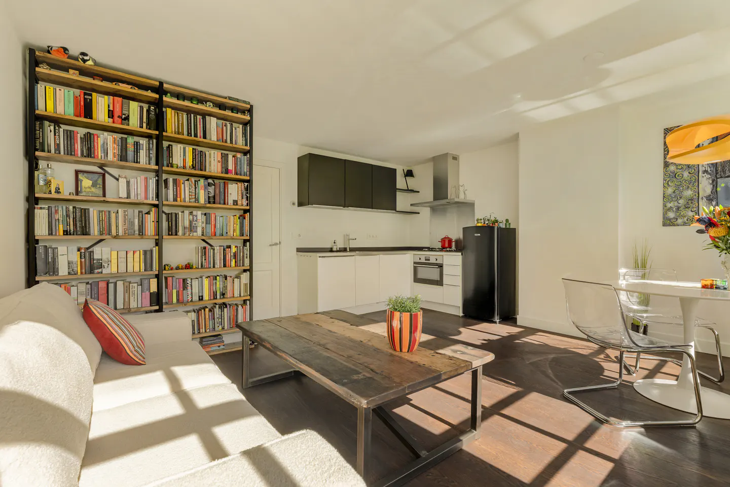 Bright apartment interior with a large bookshelf, white sofa, rustic wood coffee table, and a modern kitchen with black fridge.