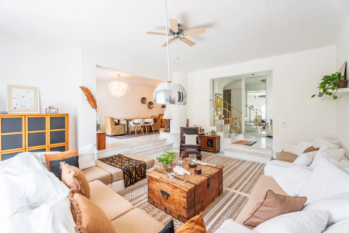Bright, multi-level living room with white walls, beige sectional sofa, and wooden trunk coffee table. Dining area and staircase visible in background.