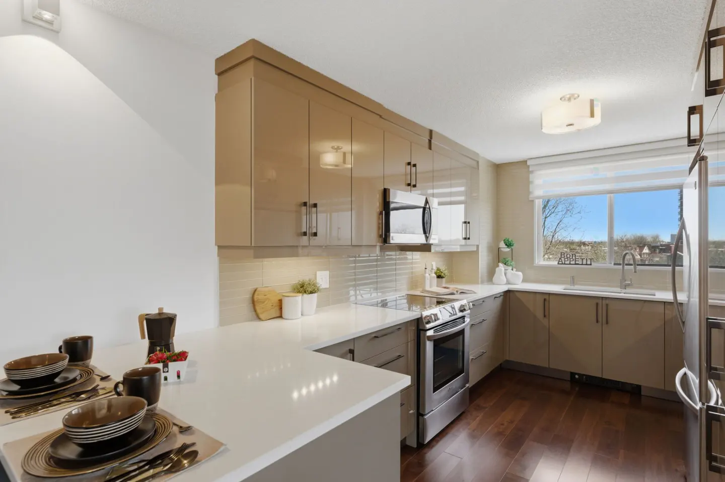 A modern kitchen with tan cabinets, stainless steel appliances, and a white countertop. A table setting is visible in the foreground.