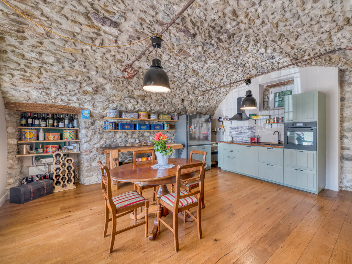 Stone-walled kitchen with wood floors, a round table with chairs, and light green cabinets. Shelves display wine and tins.