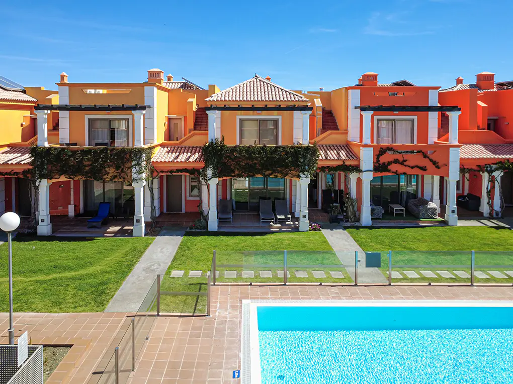 Row of orange townhouses with white trim, tile roofs, and green lawns, viewed from above a blue swimming pool.