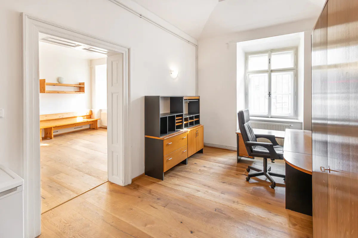 Bright room with wood floors, white walls, and a window. A desk, chair, and cabinet furnish the space. An open doorway leads to another room.