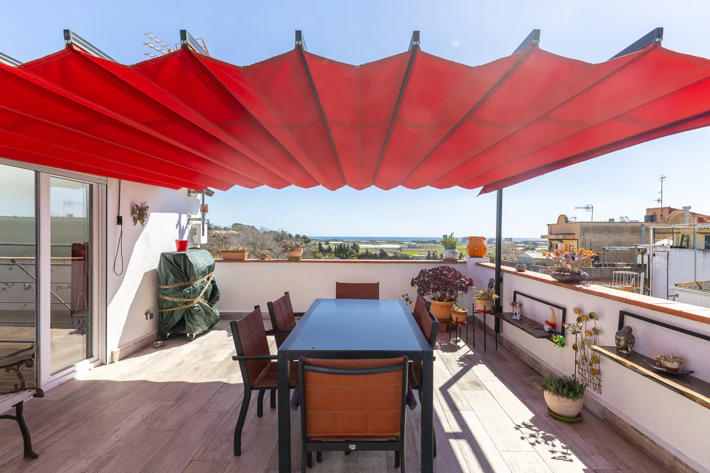 Rooftop patio with a red awning, table, and chairs. Potted plants line the white railing with a view of the landscape.