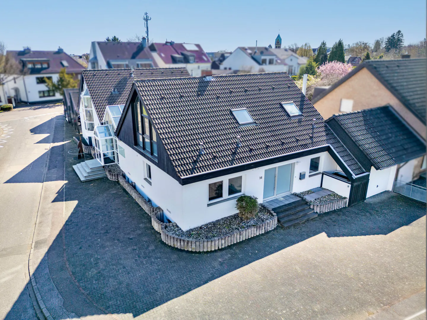 Aerial view of a white, modern house with a dark gray roof and a paved driveway. Other houses and trees are visible in the background.