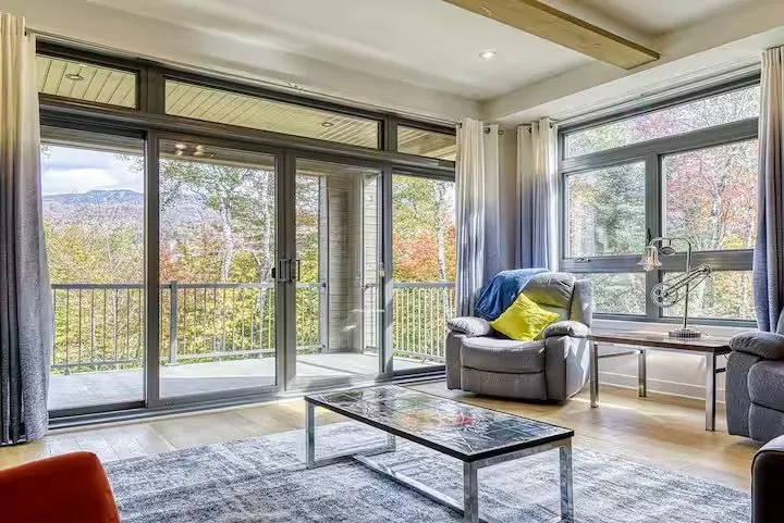 Living room with large windows showing trees and mountains. A gray recliner with a blue blanket and yellow pillow sits near a table lamp.