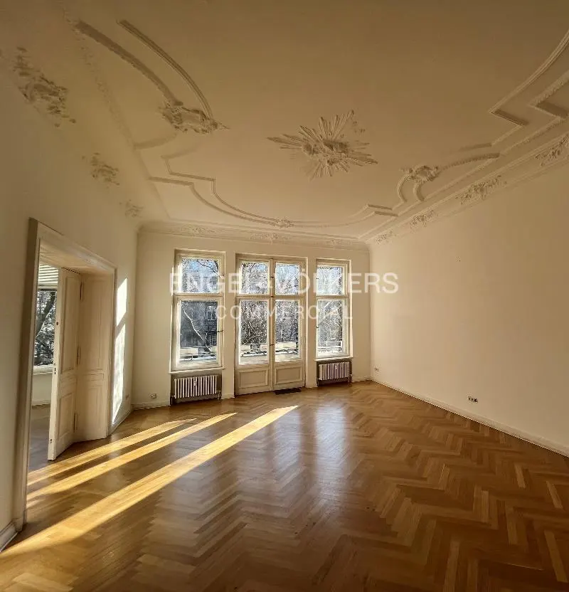 Bright, empty room with herringbone wood floors, white walls, and ornate ceiling molding. Three windows overlook trees.