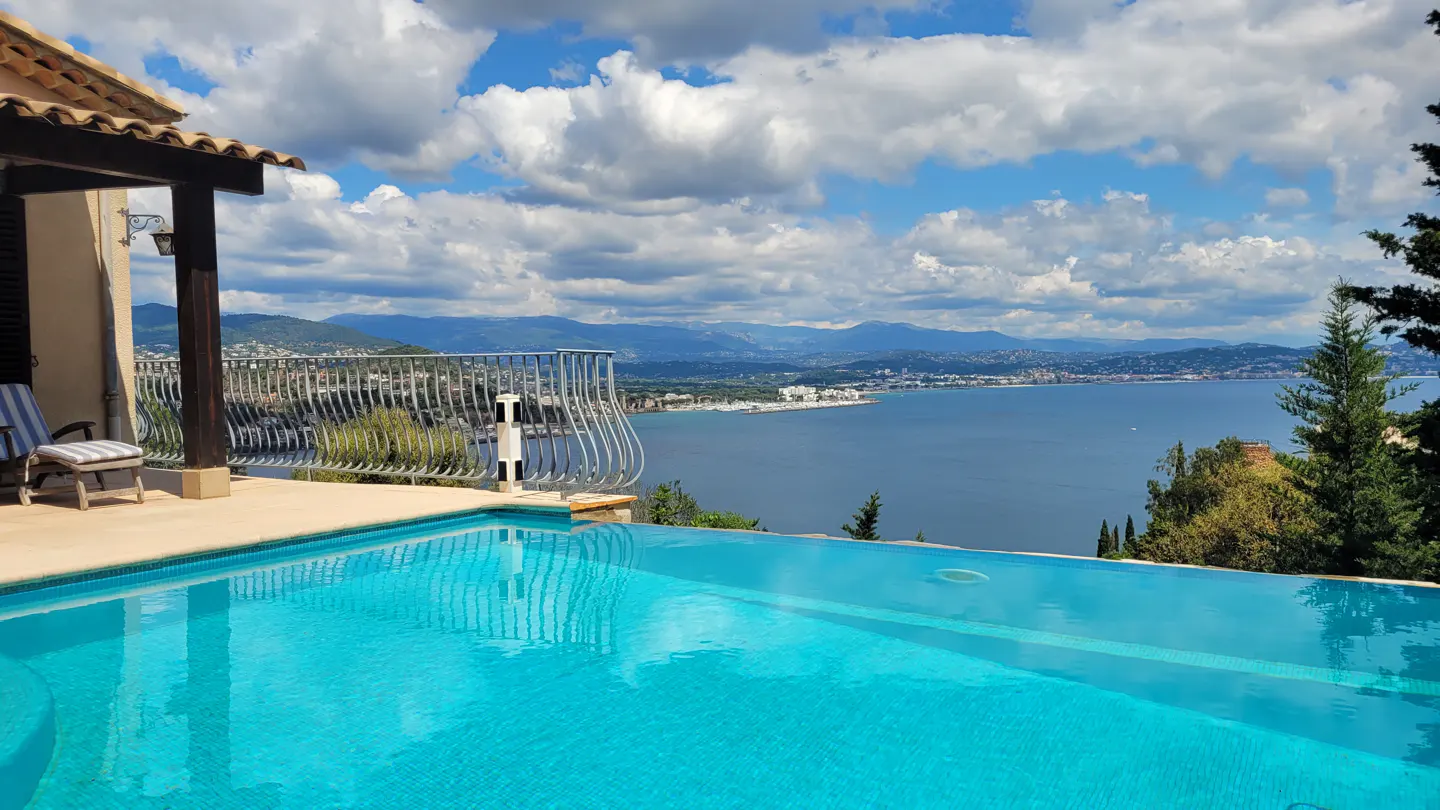 Infinity pool with blue tile overlooking the ocean, mountains, and cloudy sky. Balcony with railing and outdoor furniture.