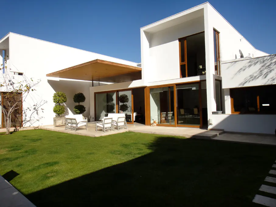 Modern white house with a green lawn, patio, and outdoor seating under a wooden awning. Large windows and sliding glass doors.