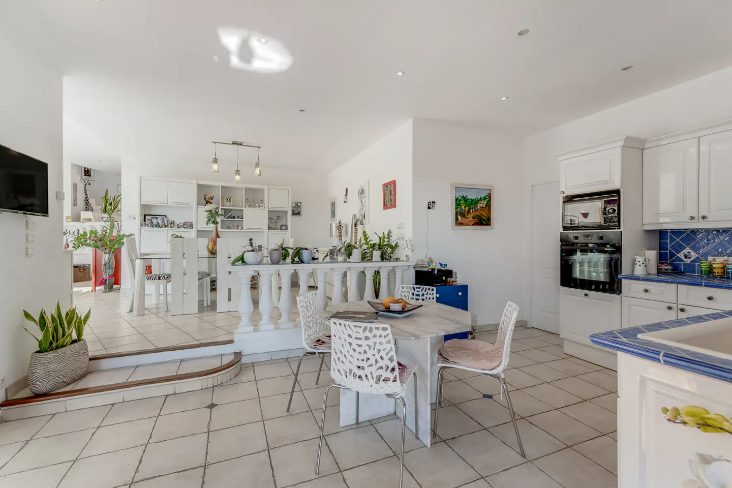 Bright, open-concept kitchen and dining area with white tile floors. A white table and chairs sit near a balustrade. White cabinets and appliances.