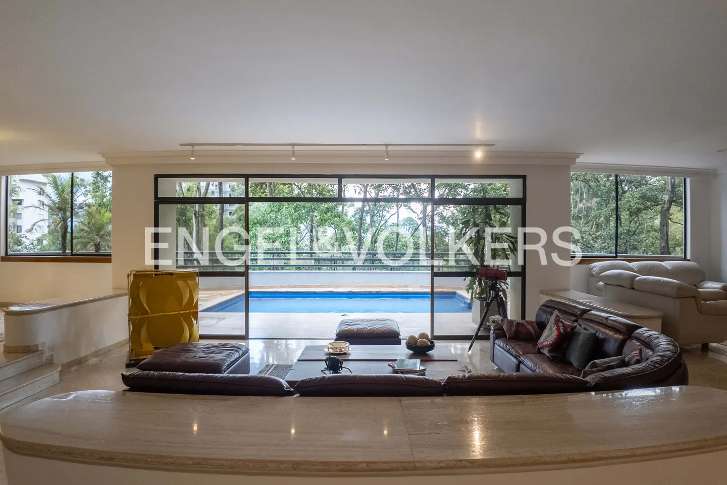 Wide shot of a living room with a brown leather sofa, a coffee table, and a view of a pool through a large window.