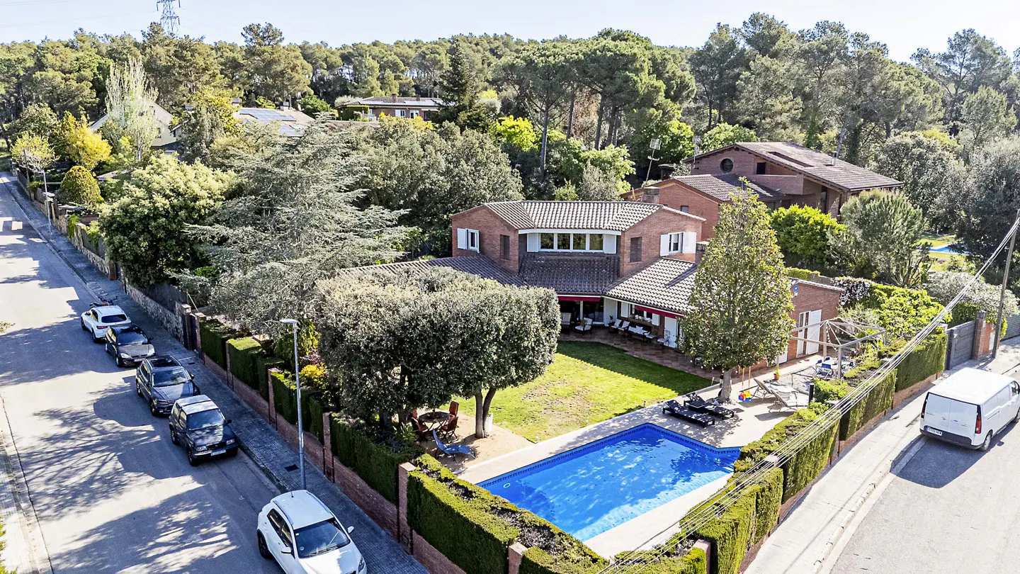 Aerial view of a two-story brick house with a blue swimming pool, green lawn, and tall trees in the background.