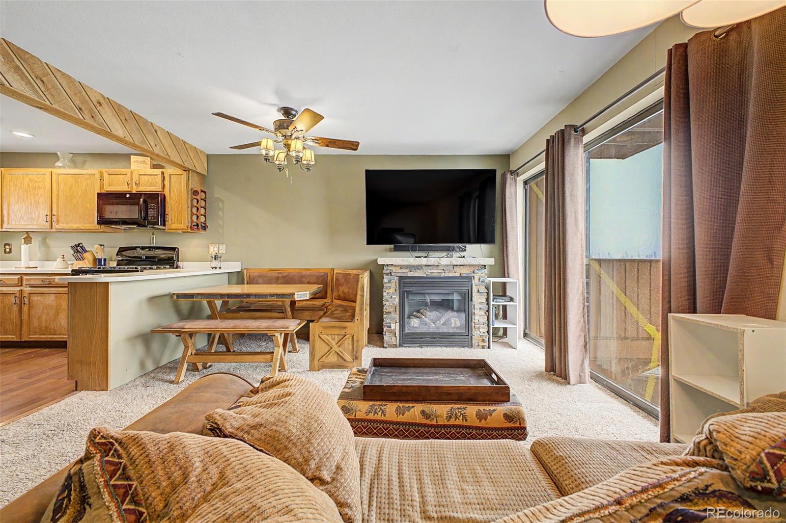 Living room with a stone fireplace, TV, wood table, and a brown sofa with patterned pillows. Sliding glass doors lead to a balcony.