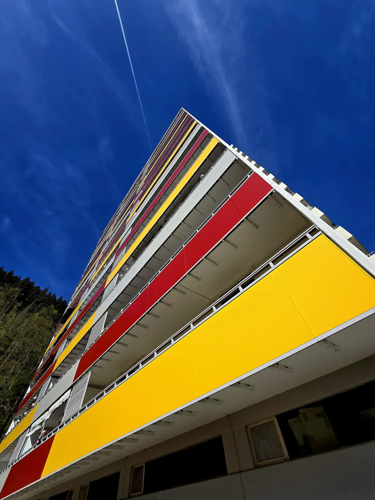 Low-angle shot of a tall building with red and yellow balconies against a bright blue sky with a contrail.