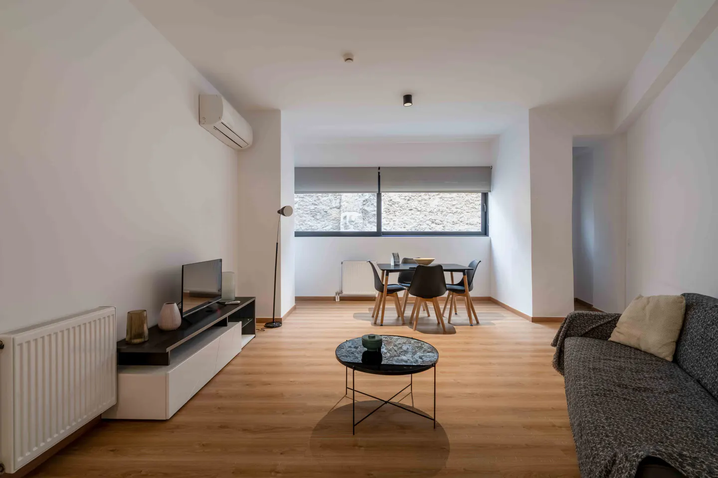 Bright living room with wood floors, white walls, and modern furniture. A dining table, sofa, TV, and coffee table are visible.