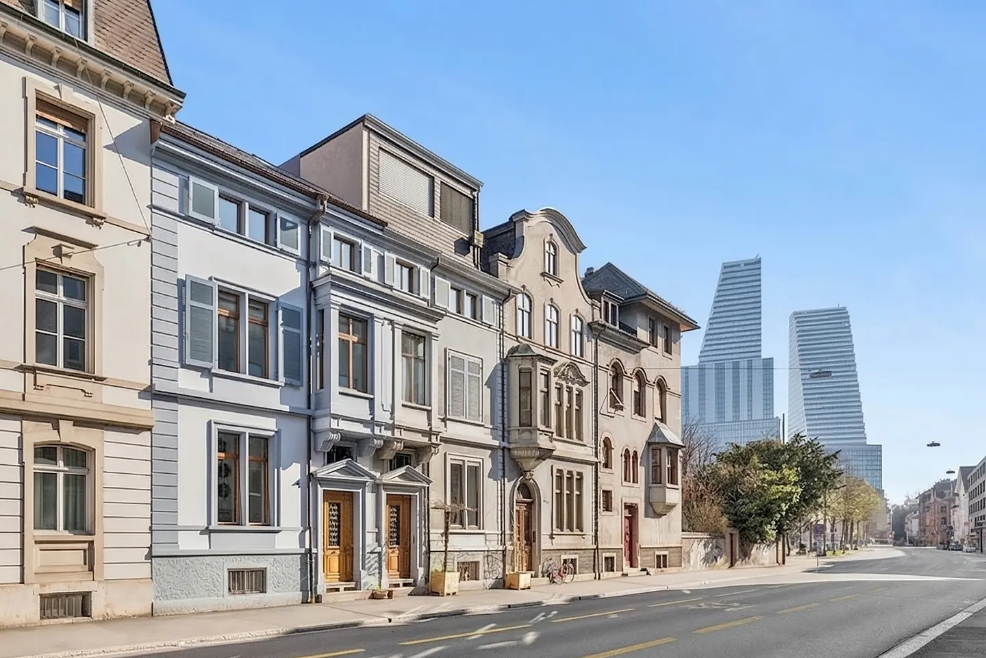 Row of European-style houses in neutral tones, with modern skyscrapers in the background under a clear blue sky.
