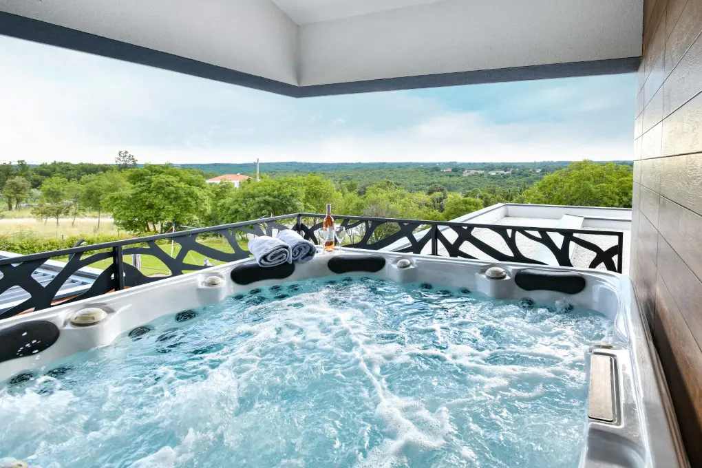 Outdoor jacuzzi with bubbling water on a balcony. Towels and wine are on the edge, with a green landscape in the background.