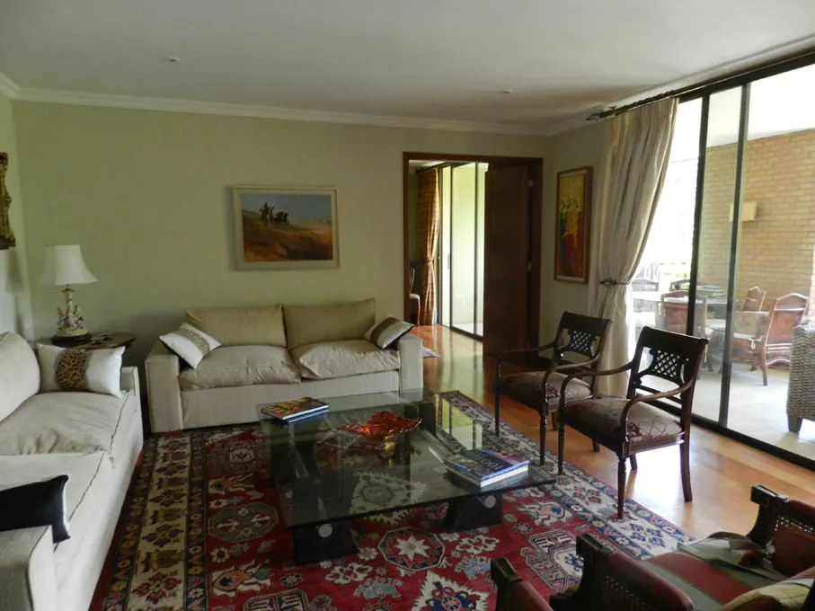 Living room with beige sofas, glass coffee table on a red patterned rug, and dark wood chairs near sliding glass doors.