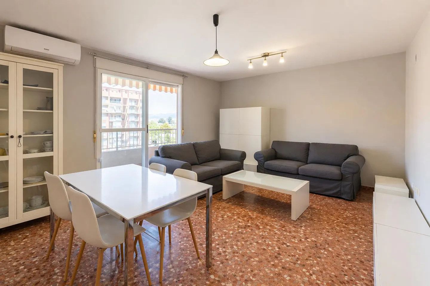 Living room with gray sofas, white table and chairs, and a cabinet. Balcony with a view in the background.