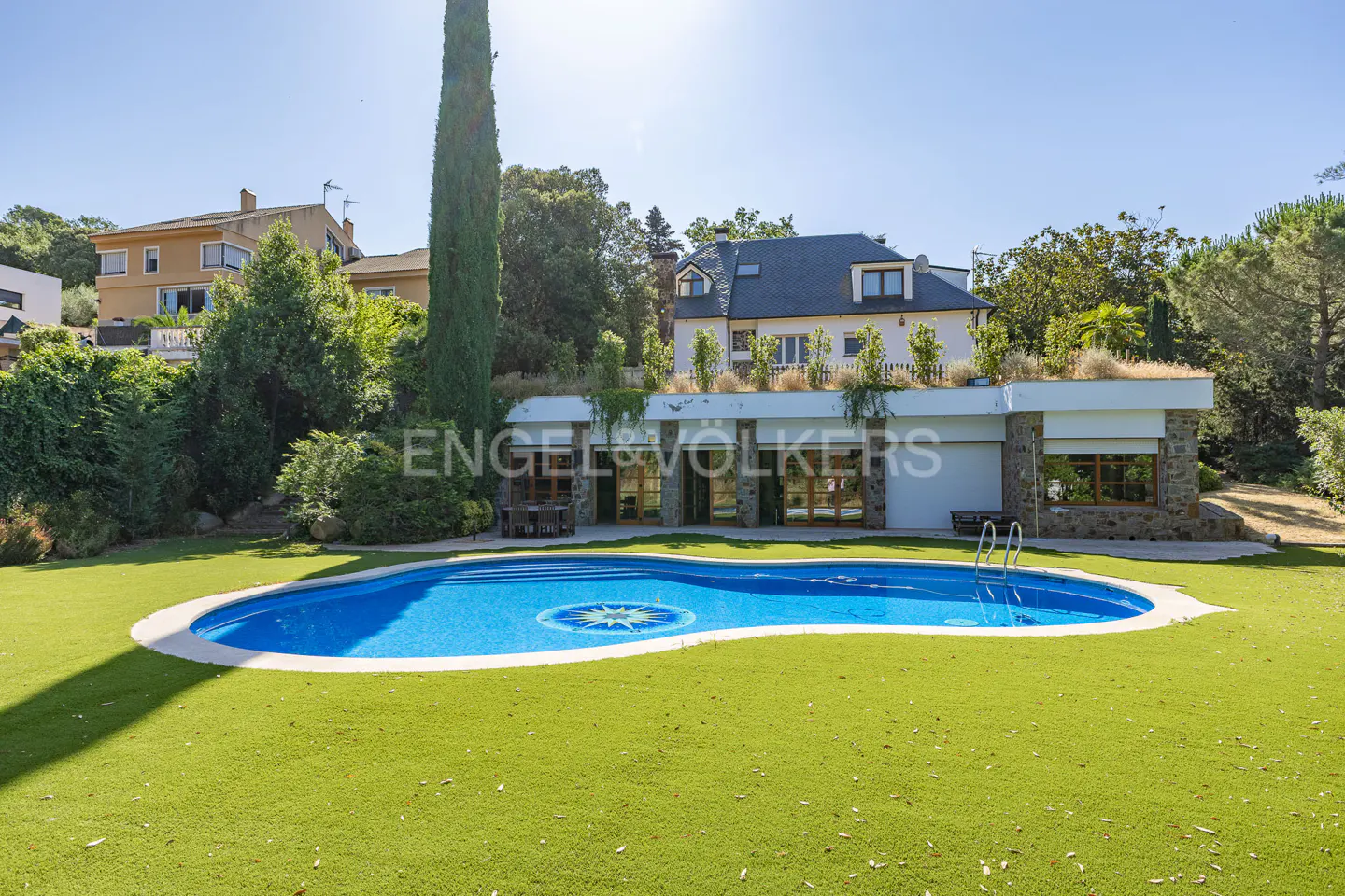 A kidney-shaped blue pool with a star design sits on green grass in front of a modern home with a green roof.