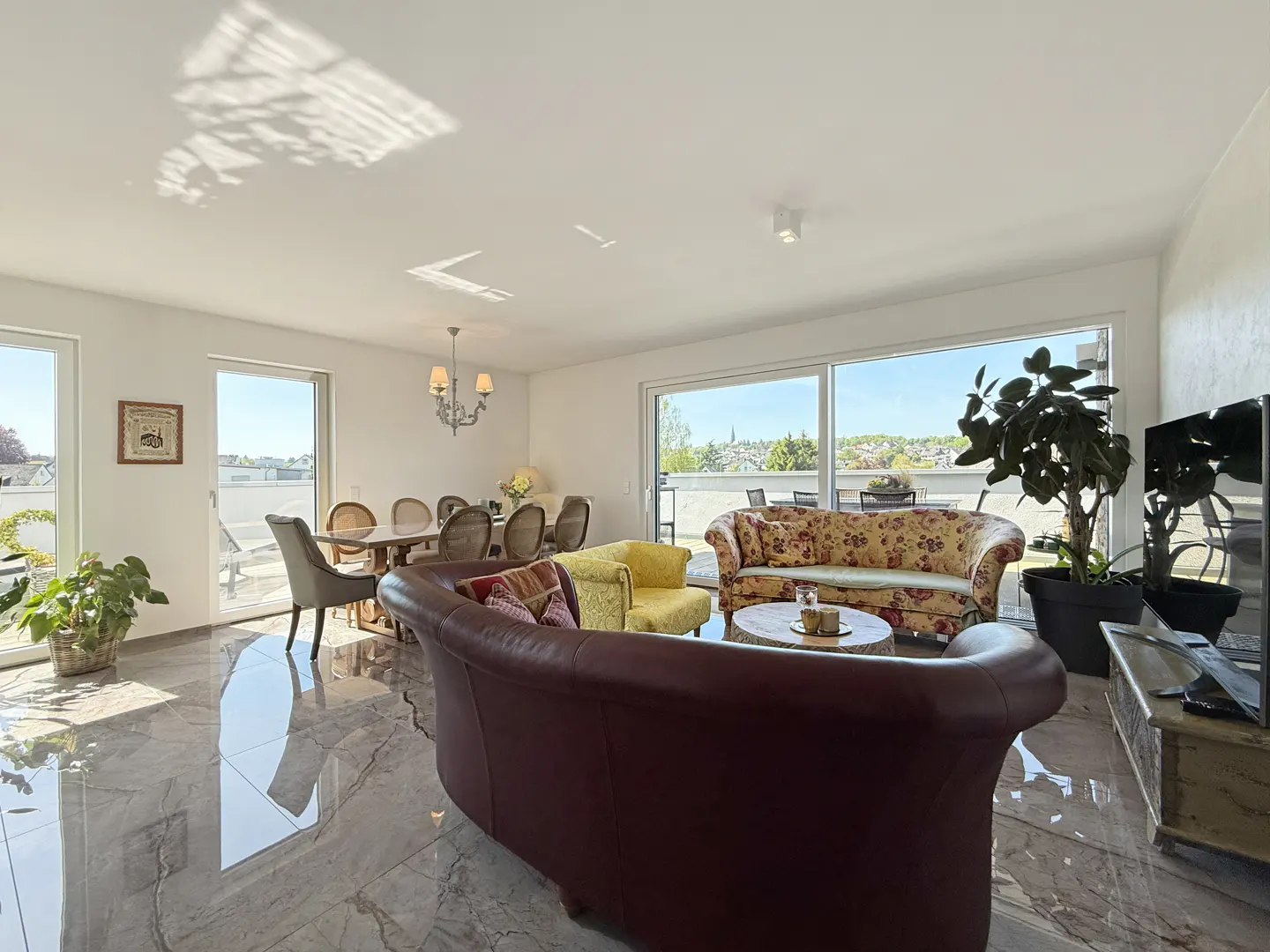 Bright, open living space with marble floors, dining table, and floral sofas. Large windows offer a view of the outside.