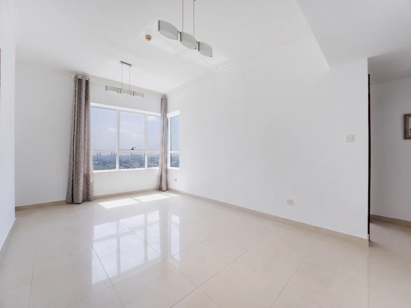 Bright, empty room with white walls, beige tile floor, and large window with gray curtains overlooking a city.