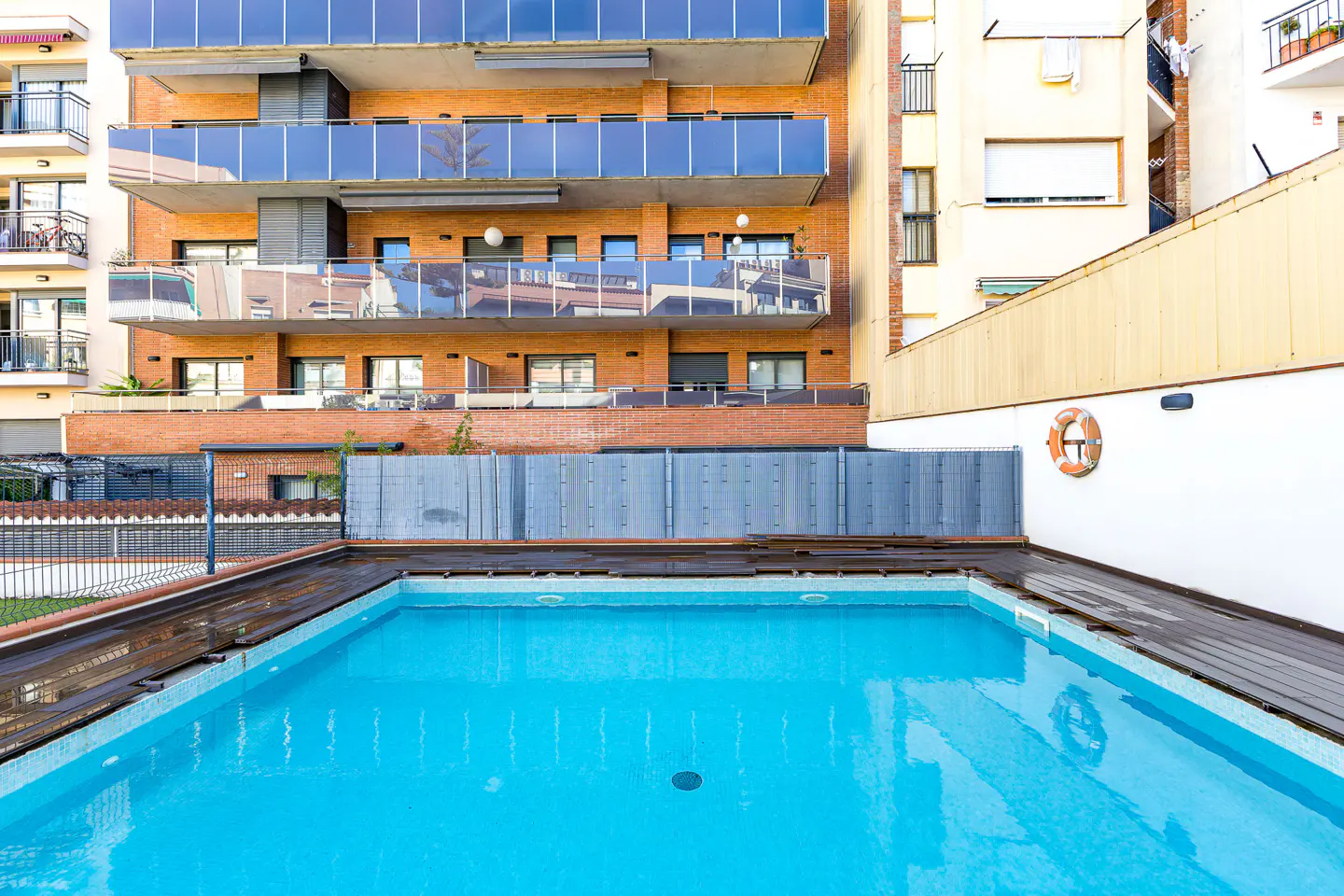 A turquoise pool is surrounded by a wooden deck and buildings. A life preserver hangs on a white wall.