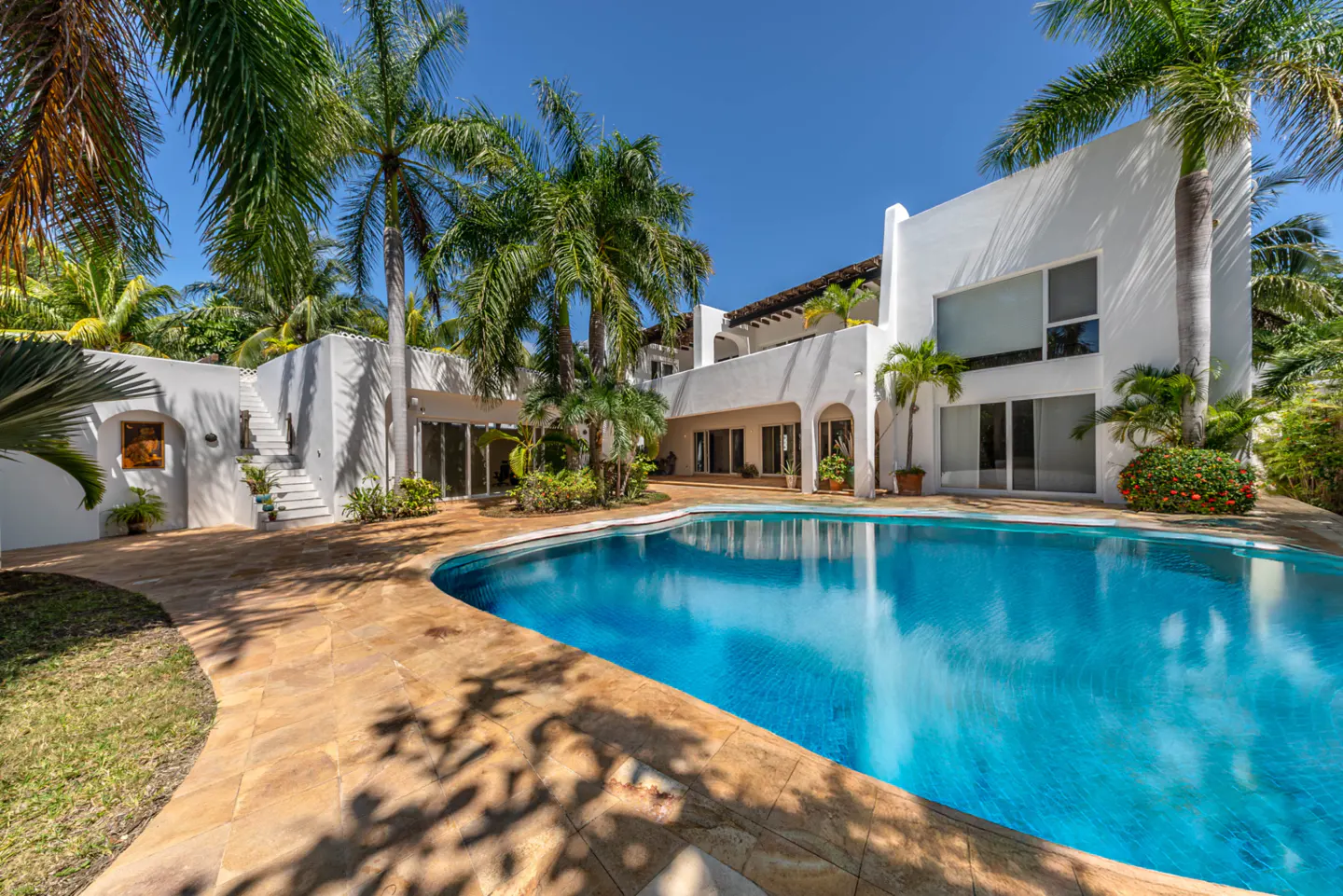 Luxury white villa with a blue pool, surrounded by palm trees under a clear blue sky.