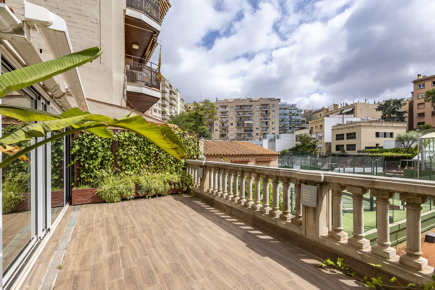 Spacious balcony with wood-look tile flooring, stone balustrade, lush greenery, and a view of tennis courts and city buildings.