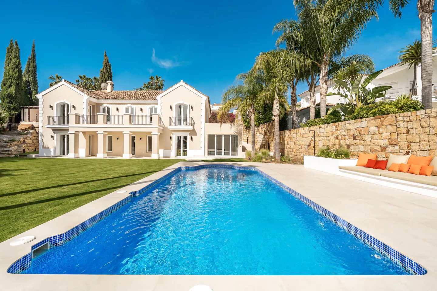 Beige two-story house with a blue tiled pool, green lawn, and palm trees under a clear blue sky. A stone wall and built-in bench with orange pillows are visible.