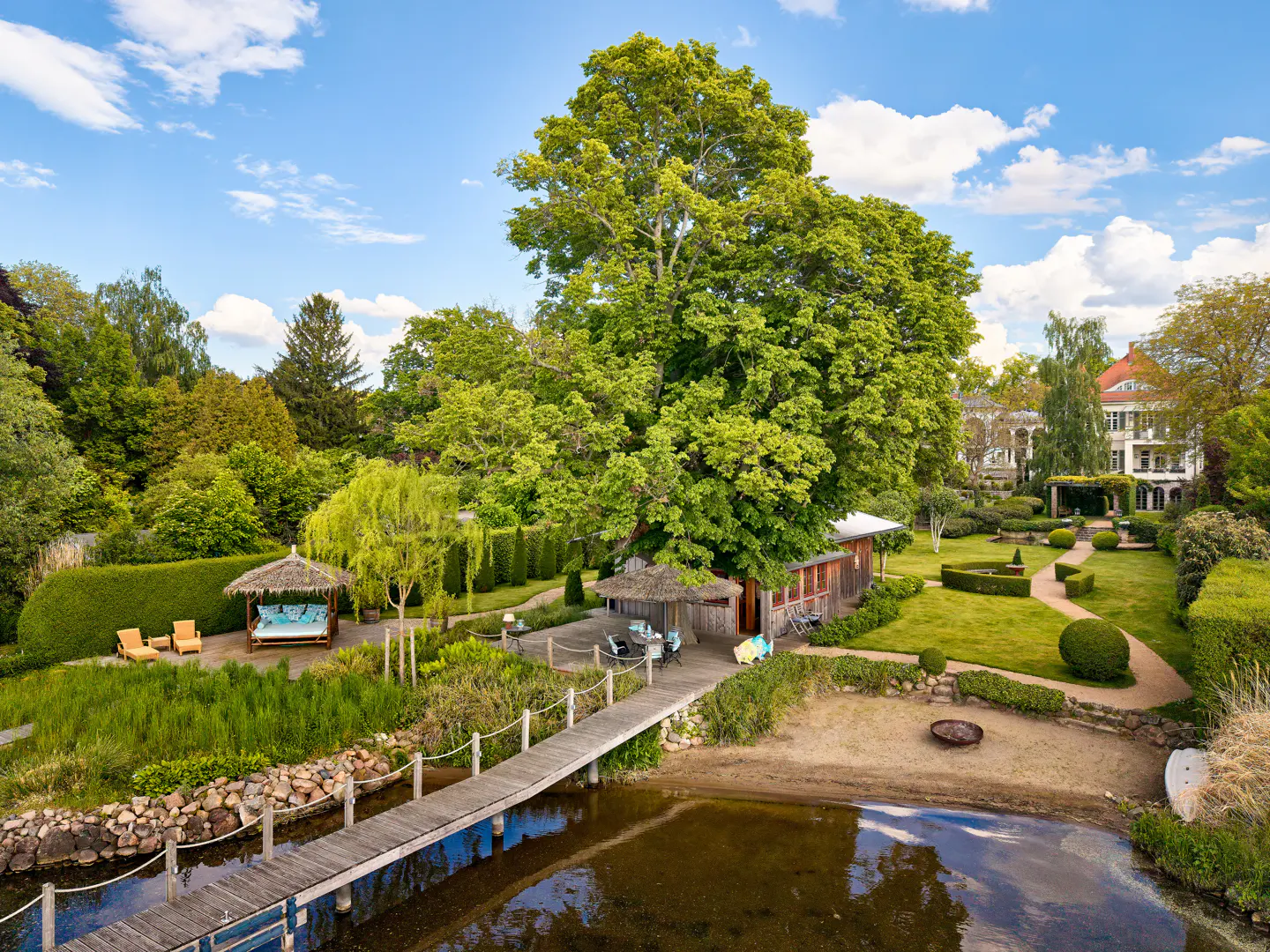 A wooden pier leads to a lush garden with a thatched-roof gazebo, a rustic cabin, and a large tree by a calm lake.