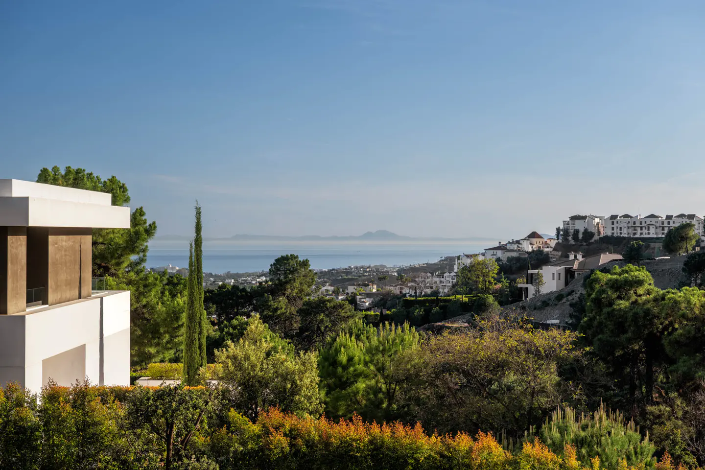 View of a white modern house overlooking a green landscape with trees, buildings, and the ocean under a blue sky.
