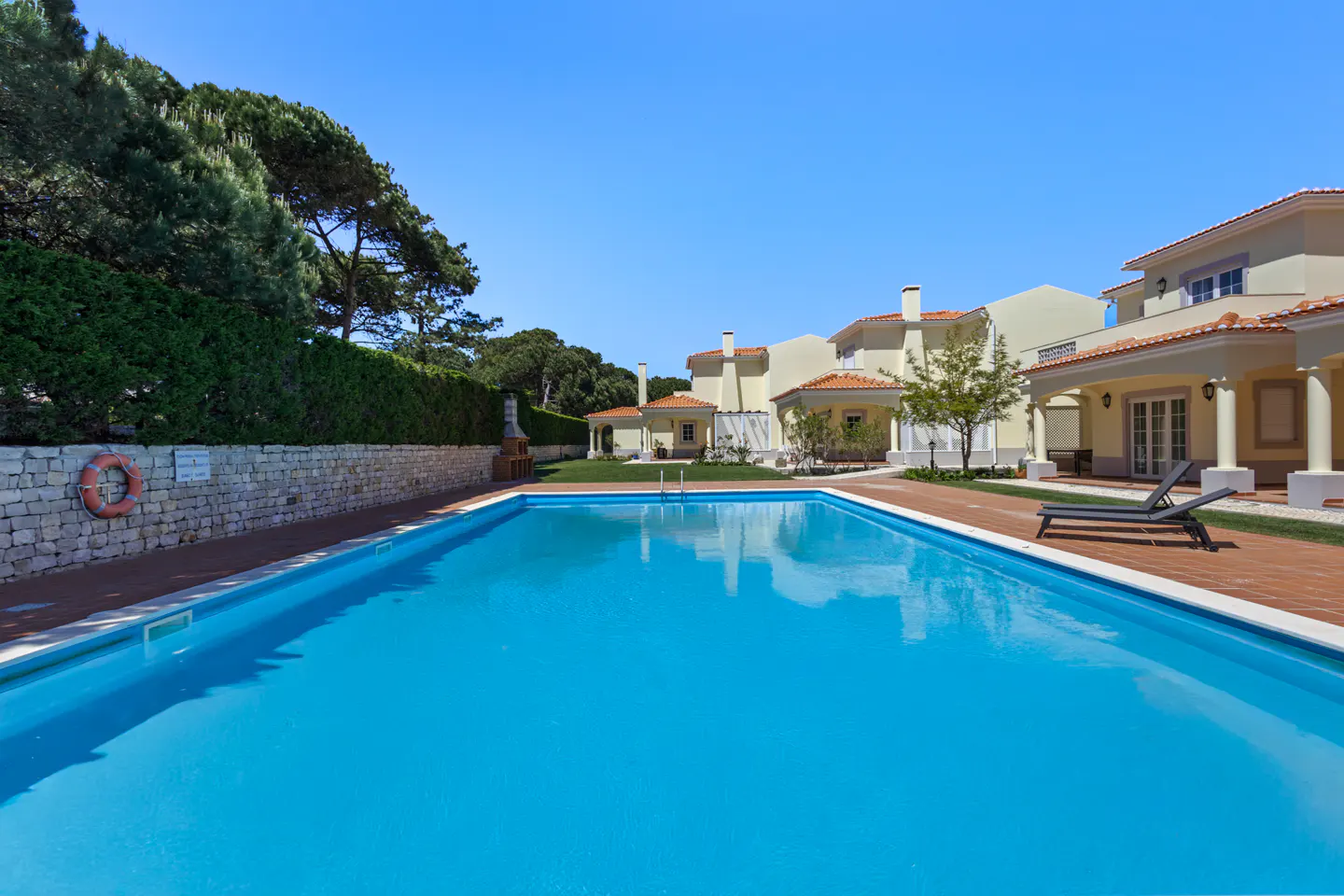 A bright blue swimming pool sits in front of a row of tan villas under a clear blue sky. A stone wall and green hedge border the pool area.
