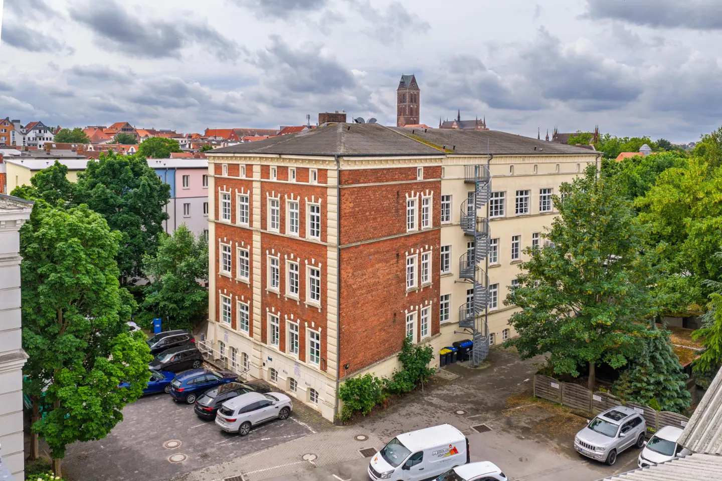 High-angle view of a multi-story brick and cream building with a spiral staircase, parking lot, and city skyline background.