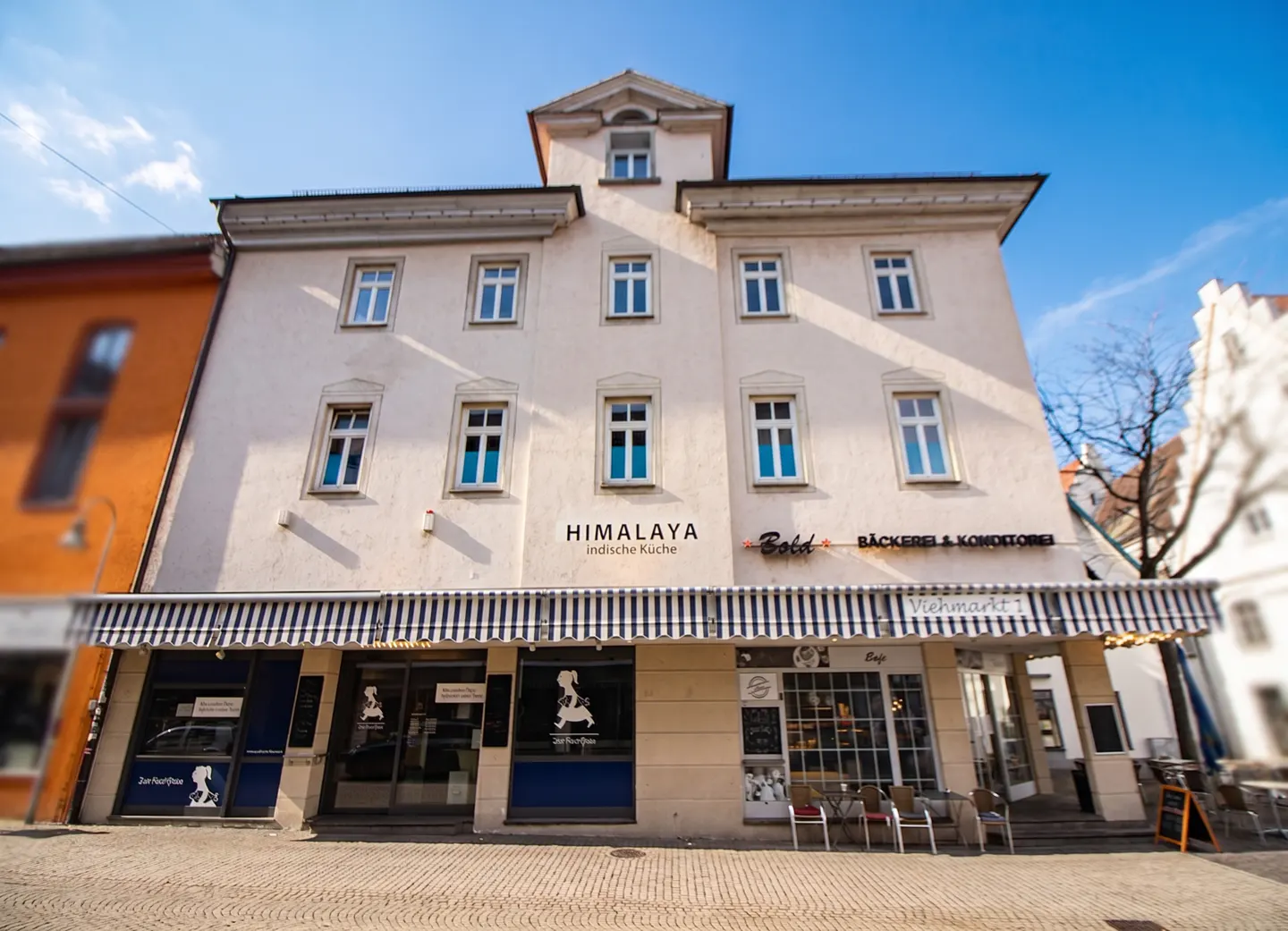 Three-story building with shops on the ground floor. "Himalaya" and "Bold" signs are visible. Blue and white striped awnings.
