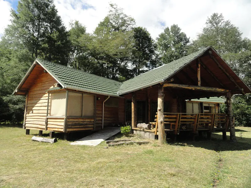 A cozy, one-story log cabin with a green roof, surrounded by lush trees and a grassy lawn.