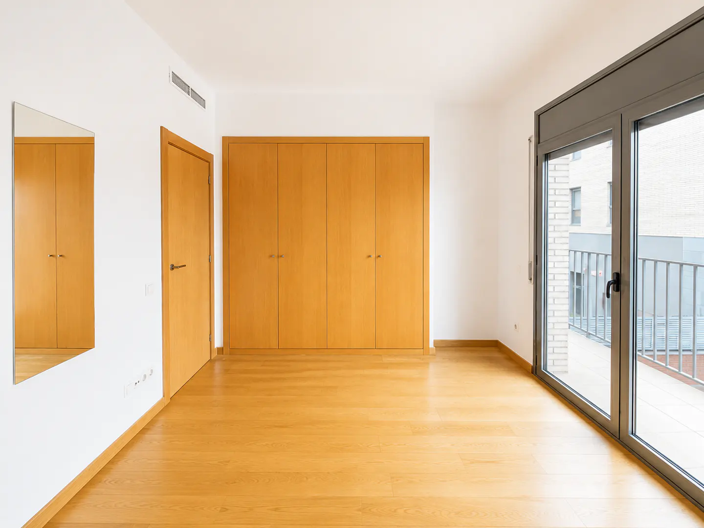 Empty room with light wood floors, large built-in wooden wardrobe, a door, a wall mirror, and a balcony with glass doors.
