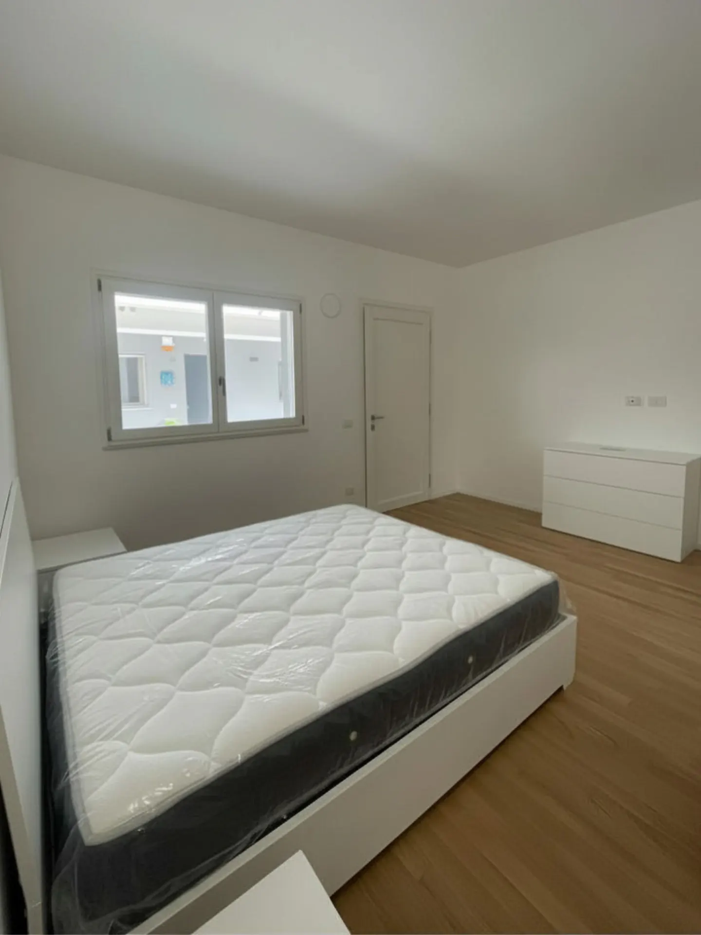 Bright, empty bedroom with a white bed frame, mattress still in plastic, wood floors, and a white dresser. A window lets in natural light.