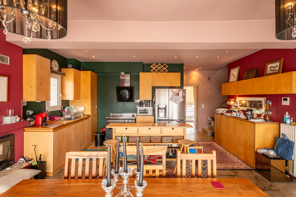 A wide shot of a kitchen and dining area with wooden cabinets, a stainless steel refrigerator, and a large wooden table with chairs. The walls are painted in red and green.