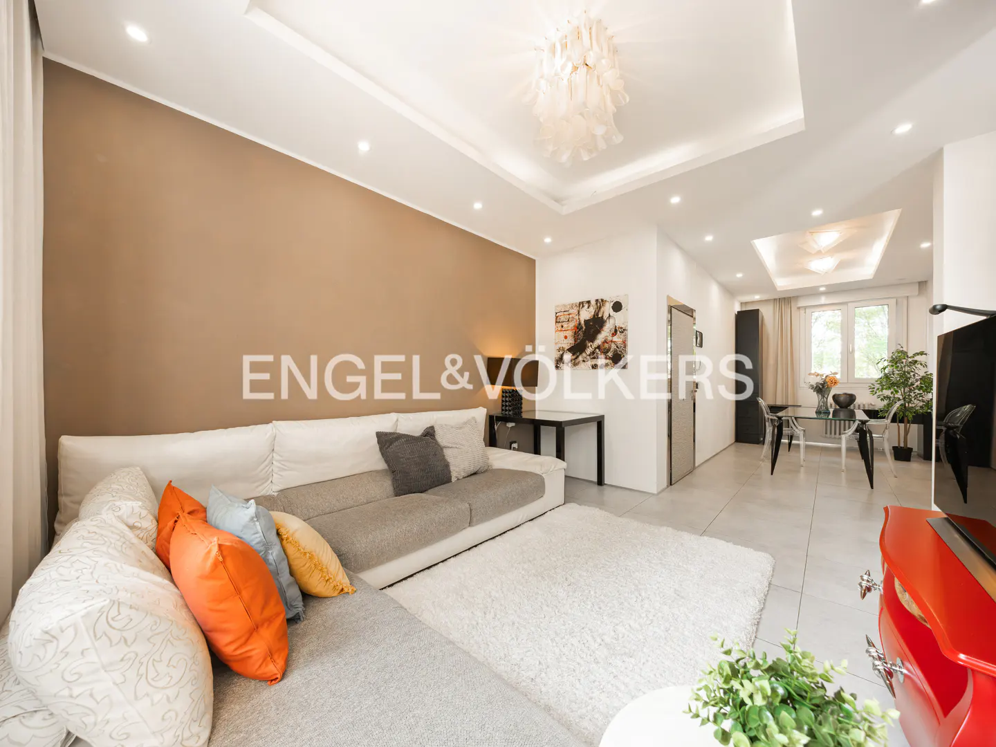 Bright, modern living room with a gray sectional sofa, colorful pillows, and a white shag rug. Dining area visible in the background.